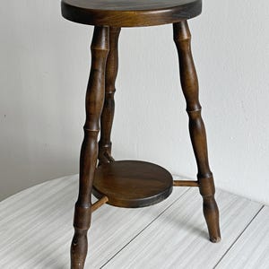 May include: A dark brown wooden plant stand featuring a circular top and a smaller circular lower shelf. The stand is supported by three turned legs with decorative spindle detailing, showcasing a rich wood grain finish and visible wear on the feet.