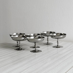 Op de afbeelding: Zes zilveren champagnecoupeglazen met een reflecterend oppervlak. De glazen hebben een ronde kom en een steel met een cirkelvormige voet. De glazen zijn gerangschikt op een lichtgekleurd oppervlak.