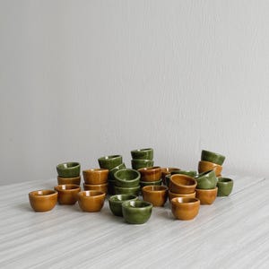 Puede incluir: Una colección de pequeños cuencos de cerámica en tonos verdes y marrones. Los cuencos están apilados en varias configuraciones, creando una exhibición visualmente atractiva.