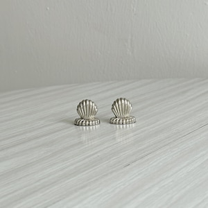 Puede incluir: Dos pequeños objetos de metal plateado con forma de conchas marinas en abanico. Cada pieza presenta una parte superior redondeada con estrías verticales y una base circular plana. Los objetos tienen un acabado pulido y reflectante.