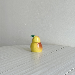 Op de afbeelding: Een keramische peer-vormige container met een deksel. De container is geel met een bruine gradiënt aan één kant en een groen blad bovenop. De container staat op een lichtgrijze ondergrond.