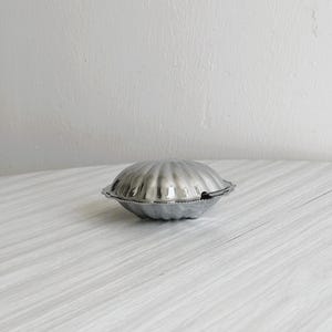 May include: A silver-colored, clam-shaped container with a textured, ridged surface. The container is closed and sits on a light-colored, wood-grain surface. The container is likely made of metal.