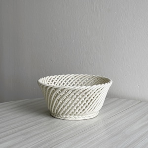 May include: A white woven basket with a round shape and a slightly flared rim. The basket is made of a thin, white material that is woven into a spiral pattern.