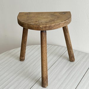 May include: A small, three-legged wooden stool featuring a semi-circular seat with a natural brown wood grain finish. The stool has three cylindrical legs attached to the underside of the flat, half-moon shaped top, showcasing visible wood texture.