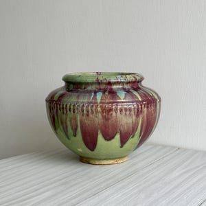 Könnte beinhalten: Eine handgefertigte Keramikvase mit einem breiten, abgerundeten Körper und einem leicht ausgestellten Rand. Die Vase zeichnet sich durch eine auffällige Farbpalette aus, mit einem grünen Sockel und einer satten burgunderfarbenen Glasur, die an den Seiten herunterläuft. Die Vase ist etwa 20 cm hoch.