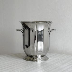 Op de afbeelding: Een gepolijste zilverkleurige metalen wijnkoeler met een geschulpte rand en twee handvatten aan de zijkanten. De emmer heeft een ronde vorm met verticale ribbels en een ronde voet, waarbij het licht reflecteert op het gladde metalen oppervlak.