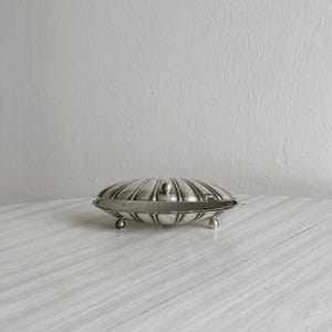 May include: A silver-colored, clam-shaped trinket box with a hinged lid and small ball feet. The box has a ribbed design and is displayed on a light-colored, wood-grain surface. The background is a plain, light gray wall.