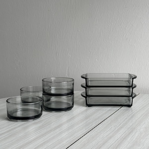 Puede incluir: Conjunto de cuatro cuencos de vidrio transparente con bordes negros, apilados en dos grupos. Los cuencos están sobre una superficie de madera clara. También hay tres fuentes rectangulares de vidrio transparente con bordes negros, apiladas una encima de la otra.