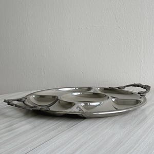 Peut inclure: Un plateau de service rond en métal argenté doté de cinq compartiments distincts, comprenant une section circulaire centrale entourée de quatre segments incurvés. Le plateau possède deux poignées ornées et texturées aux finitions sombres.