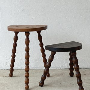 May include: Two wooden stools with semi-circular seats and three legs each. The legs feature a distinct bobbin or spool-turned design with rounded segments. The taller stool is a medium brown, while the shorter stool is a darker, espresso-toned wood.
