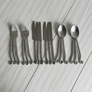 Può includere: Un set di posate in acciaio inossidabile, tra cui forchette, coltelli e cucchiai. I manici hanno un design arricciato unico. Le posate sono disposte su una superficie di legno chiaro.