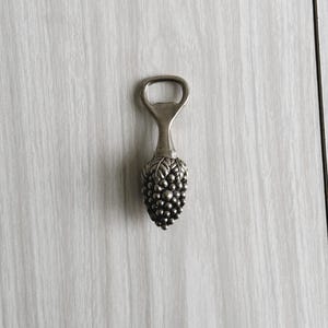 May include: Silver-toned bottle opener with a grape cluster design. The opener features a loop for leverage and a detailed grape cluster handle. A decorative and functional barware item.
