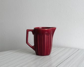 May include: A red ceramic pitcher with vertical ridges. The pitcher has a handle and a spout.