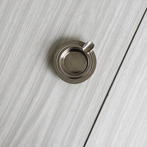 May include: A small, round, silver ashtray with a decorative beaded edge and a built-in rest for a cigarette. The ashtray is made of metal and sits on a white, textured surface.