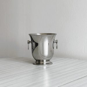 Puede incluir: Una cubitera de vino plateada pulida con dos asas de anillo. La cubitera tiene una parte superior ancha y abierta y una base decorativa. La superficie reflectante de la cubitera crea una apariencia brillante y elegante. Perfecto para enfriar vino o champán.