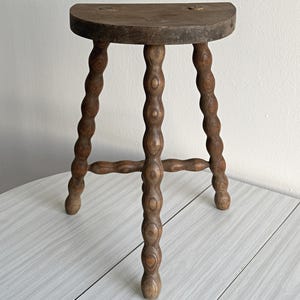 May include: A three-legged wooden stool with a semi-circular seat and turned, bobbin-style legs. The wood features a dark brown finish with visible grain patterns. The legs are connected by horizontal stretchers, creating a stable base structure.
