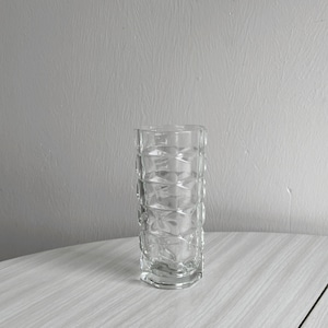 Puede incluir: Un jarrón de vidrio transparente con un patrón geométrico. El jarrón está vacío y se encuentra sobre una superficie de madera clara.