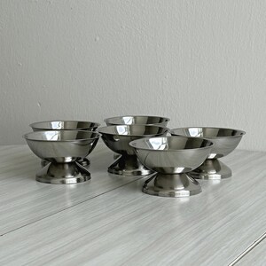 May include: A set of five polished stainless steel dessert bowls, each featuring a wide, shallow basin and a sturdy, flared pedestal base. The bowls have a smooth, reflective metallic finish and are arranged in a cluster on a light surface.