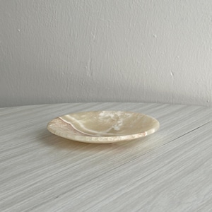 Peut inclure: Un plat circulaire peu profond fabriqué en pierre polie avec un aspect beige crème translucide. La surface présente des bandes naturelles tourbillonnantes de tons plus clairs et plus foncés, créant une texture organique lisse et brillante.
