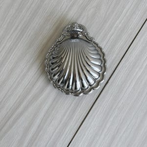 Könnte beinhalten: Eine silberne Schale in Muschelform mit einer dekorativen Krone auf der Oberseite.