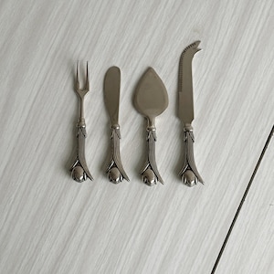 Puede incluir: Juego de cuatro cuchillos y utensilios para servir queso en tono plateado. Incluye un tenedor de dos puntas, un cuchillo para mantequilla, una espátula para queso y un cuchillo para queso con hoja curva. Cada utensilio tiene un mango decorativo con diseño floral.