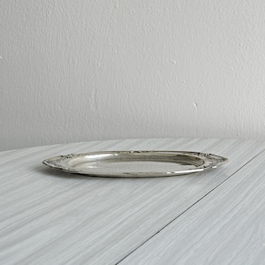 Peut inclure: Un plateau de service argenté vintage de forme ovale allongée. Le plateau présente un motif floral décoratif sur le bord. La surface réfléchissante du plateau est visible, et il repose sur une surface de couleur claire.
