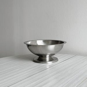 Puede incluir: Un cuenco de pedestal de acero inoxidable con una base ancha y poco profunda y un borde decorativo con cuentas. El cuenco se asienta sobre una base de pedestal redonda y corta. El cuenco se muestra sobre una superficie clara y texturizada.