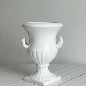 May include: A glossy white ceramic urn-style vase featuring a wide flared rim, two small curved side handles, and a fluted body that tapers down to a pedestal base. The base is square-shaped with vertical ridges extending from the bottom.