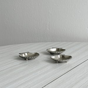 Peut inclure: Trois petits plats en métal argenté en forme de coquilles Saint-Jacques. Chaque plat présente une texture nervurée sur l'extérieur, un bassin intérieur lisse et est soutenu par trois minuscules pieds arrondis qui surélèvent la base.