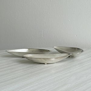Peut inclure: Trois plats en métal argenté en forme de bateau avec des bords lisses et incurvés et de petits pieds. Les récipients présentent un design allongé et effilé avec un bassin intérieur peu profond, disposés pour montrer leur forme.