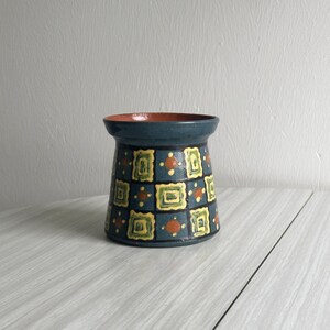Op de afbeelding: Een kleine, blauwe keramische vaas met een geometrisch patroon in geel, groen en oranje. De vaas heeft een uitlopende bovenkant en een ronde bodem.