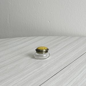 May include: A small, clear glass jar with a yellow cushion top and a silver-colored metal rim. The jar sits on a light gray wooden surface. The jar is likely a vintage sewing accessory.