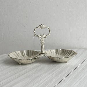 Può includere: Piatto da portata argentato a due scomparti con manico decorativo. Il piatto ha un bordo smerlato e poggia su piccoli piedini. Il manico presenta dettagli ornati. Un pezzo in stile vintage per servire snack o condimenti.