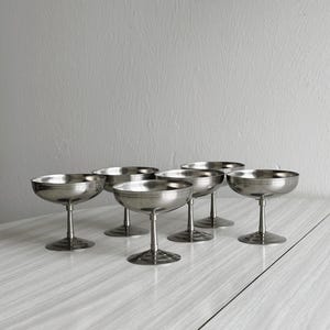 Peut inclure: Six coupes à champagne en argent avec un motif cannelé sur la base. Les coupes sont disposées en rangée sur une surface en bois clair.