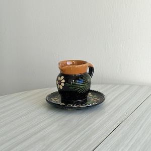 May include: A small, brown ceramic pitcher with a floral design. The pitcher has a handle and a matching saucer. The pitcher and saucer are decorated with white and green flowers.