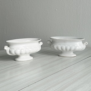 May include: Two white ceramic vessels with an oval shape, featuring a fluted body design, a pedestal base, and small scroll-shaped handles on each side. The bowls have a smooth, glossy finish and are positioned side by side on a flat surface.