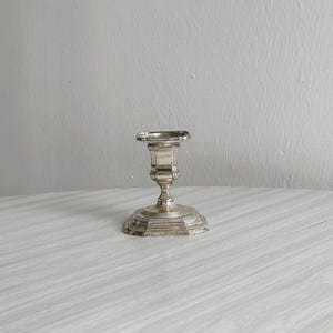 May include: A silver candlestick holder with a round base and a detailed, ornate design. The candlestick has a short stem and a small cup to hold a candle. The piece is made of a shiny, reflective metal.