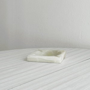 May include: A square, off-white onyx ashtray with a shallow, recessed bowl. The ashtray has a smooth, polished surface and a subtle, translucent quality. The edges are clean and defined, and the overall design is minimalist.