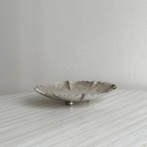 Peut inclure: Un bol décoratif argenté avec un bord festonné et un motif floral. Le bol repose sur trois petits pieds. Le bol est vide et repose sur une surface claire à grain de bois.