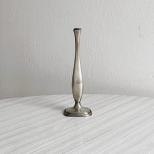 Puede incluir: Un candelero plateado con un diseño esbelto y alargado. La base es redonda con un borde de cuentas, y la parte superior es ligeramente acampanada. El candelero se muestra sobre una superficie blanca y gris.