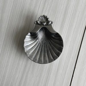 May include: A silver-colored, seashell-shaped dish with a detailed, ridged design. The dish has a decorative, fan-shaped accent at the top. The dish is likely made of metal and is suitable for holding small items.
