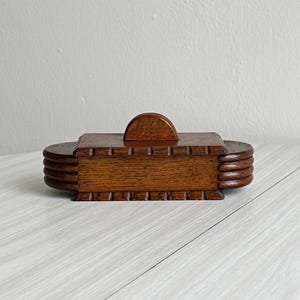 Può includere: Una scatola di legno vintage con coperchio e manico a forma di semicerchio. La scatola è rettangolare con estremità arrotondate e una serie di scanalature orizzontali. Il legno ha un caldo tono marrone, che suggerisce un oggetto antico o artigianale.
