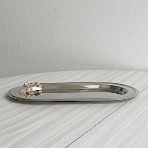 Puede incluir: Una bandeja ovalada plateada con un adorno decorativo dorado. La bandeja está vacía y descansa sobre una superficie clara con vetas de madera. La bandeja mide aproximadamente 30 cm de largo.