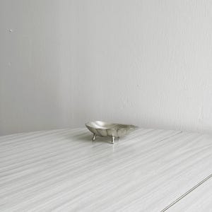Può includere: Un piccolo piatto argentato con piedini e bordo ondulato. Il piatto è su una superficie di legno bianco.
