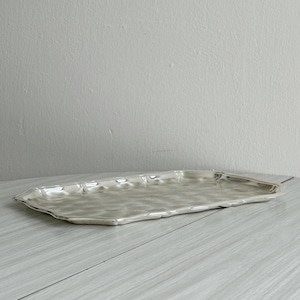 May include: A rectangular silver-toned serving tray with a polished, reflective finish and a distinctively hammered, undulating surface texture. The tray features slightly raised, scalloped edges that create a soft, flowing border around the perimeter.