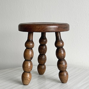 May include: A small, round wooden stool featuring a dark brown finish and a smooth, circular seat. The stool is supported by three legs, each constructed from a series of four stacked, rounded wooden spheres that taper toward the base.