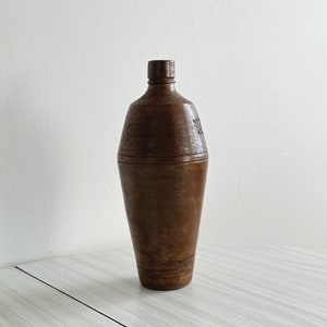 Può includere: Una bottiglia di ceramica marrone con fondo arrotondato e collo stretto. La bottiglia ha una superficie testurizzata e sembra essere vintage.