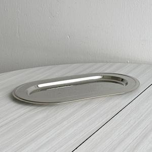 Könnte beinhalten: Ein poliertes, ovales Serviertablett mit erhöhtem Rand. Das Tablett besteht aus glänzendem, silberfarbenem Metall und reflektiert das Licht. Das Design ist schlicht und elegant, geeignet zum Servieren von Speisen oder zum Präsentieren von Gegenständen.