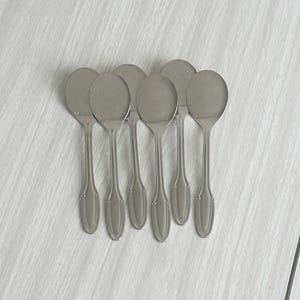 Puede incluir: Un juego de seis cucharas de acero inoxidable con cuencos ovalados y mangos decorativos. Las cucharas están dispuestas en fila sobre una superficie clara. Cada cuchara mide aproximadamente 15 cm de largo.