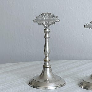 May include: A silver-toned metal stand featuring a slender, tapered stem and a wide, circular base. The top of the stand is finished with a decorative, fan-shaped crest that has a horizontal bar, providing a stable support structure for display.
